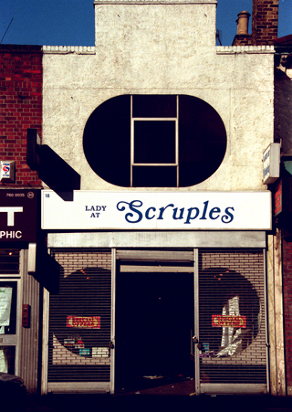 Sign above shop reads: “Lady at Scruples”. A narrow terraced two-storey shopfront. A striking oval window is set into the first floor, and the roof profile features a large sticking-up rectangular detail. The shopfront on the ground floor has an open door-shaped space in the middle, with grid-style shutters drawn down on either side. A sign above reads “Lady at Scruples”, with the “Lady at” part in smaller text than the “Scruples”.
