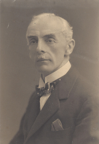 A black-and-white portrait photo of a thin-to-average white man in his late 60s or early 70s, with receding white hair and a toothbrush moustache.  He is wearing a white collared shirt, a bowtie, and a suit jacket with a pocket square.