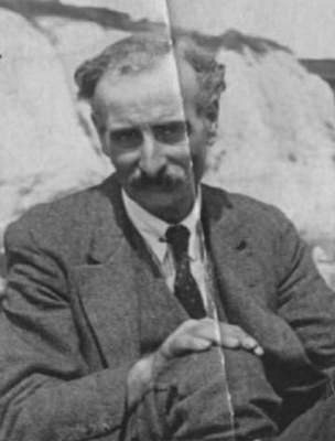 A black-and-white photo of a white man with receding wavy hair and a relatively substantial moustache.  He is wearing a suit with waistcoat and a tie, and appears to be sitting on the ground with a hand on one knee.  In the background is something that could be a rock or cliff.
