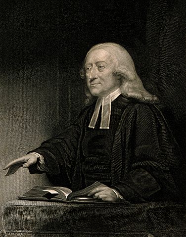 A monochrome picture of a white man wearing a dark cassock; he is reaching out one hand above a book.